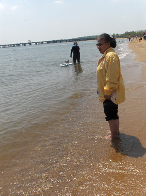 2 photos show Nanta still standing in the water.  A man is standing deeper in the water and talking with her.  He is wearing a 'wet suit' (covering his body for warmth) and the water is above his knees.  He wears headphones *to hear the metal detector) and has a metal detector with a hose, and a little floating raft to hold his treasure.